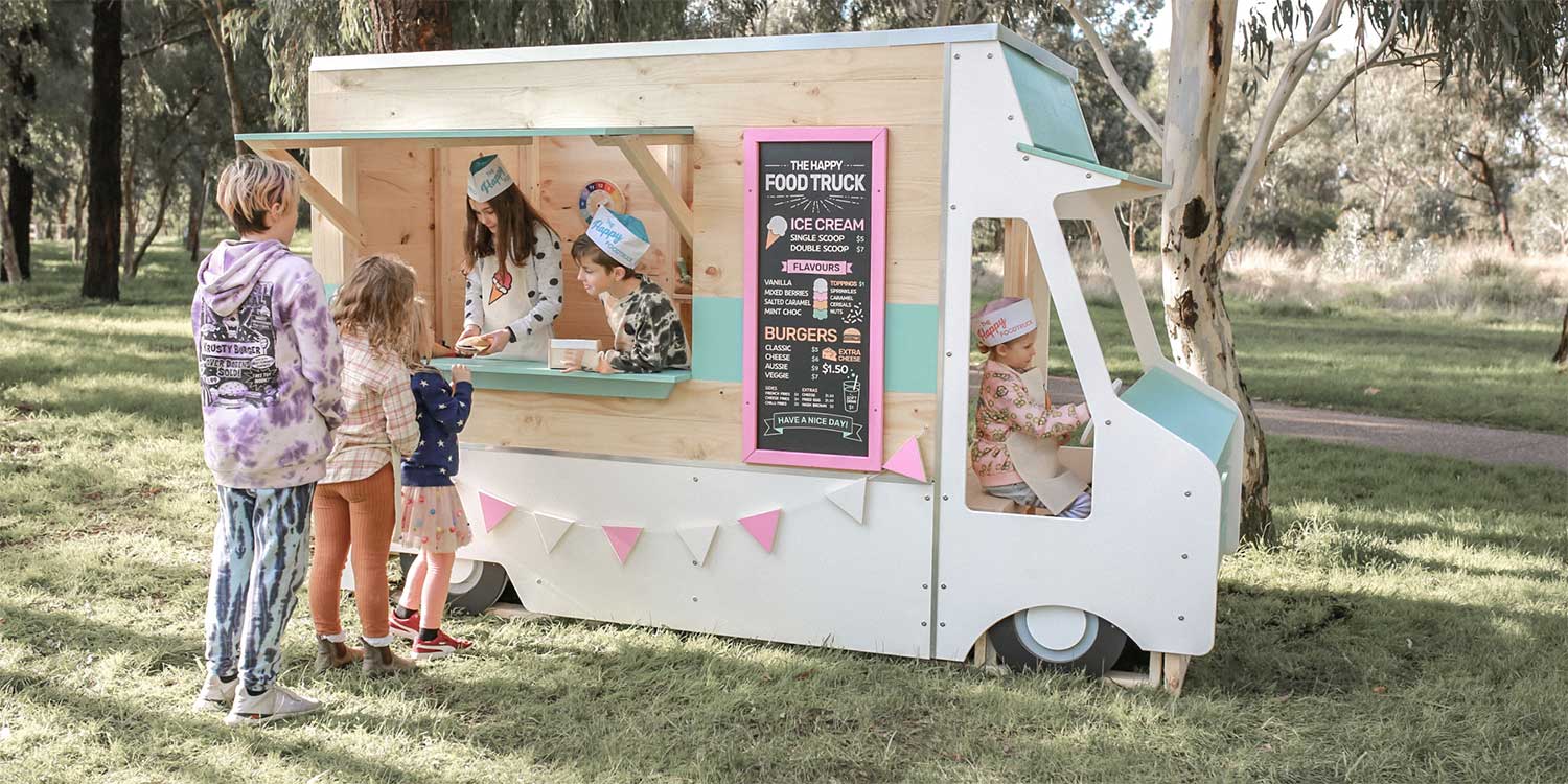 A wooden cubby house styled like a food truck for kids to play in. The image shows kids serving food to the customers lined up and a child driving the food truck.