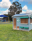 A vet themed grey and green cubby house on grass in a school yard with play equipment in the background