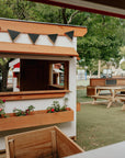A white painted wooden cubby house on school grounds with a mud kitchen and picnic tables