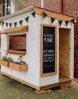 A bright white painted cafe cubby house with stained timber accents and chalkboard artwork in a school yard