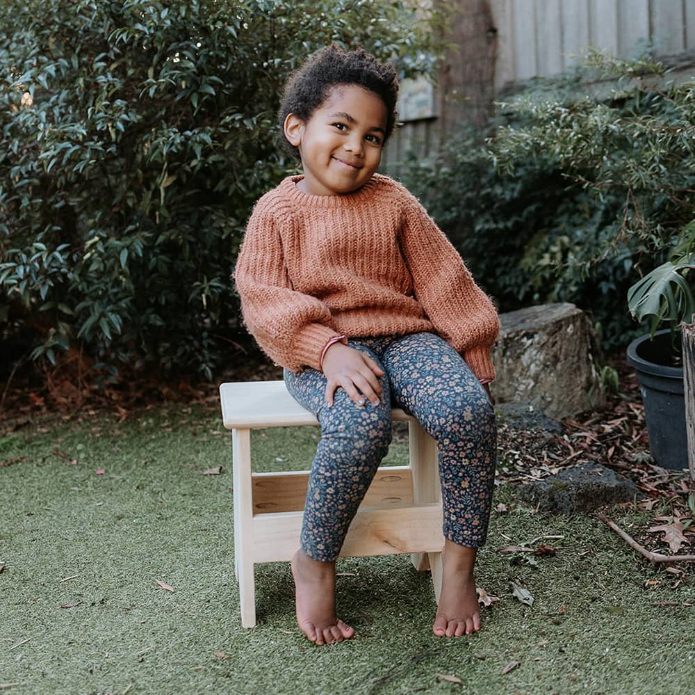 A girl sits happily on a wooden carpenter stool