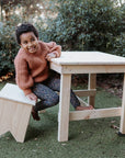 A girl sits at a table on a pine carpenter stool