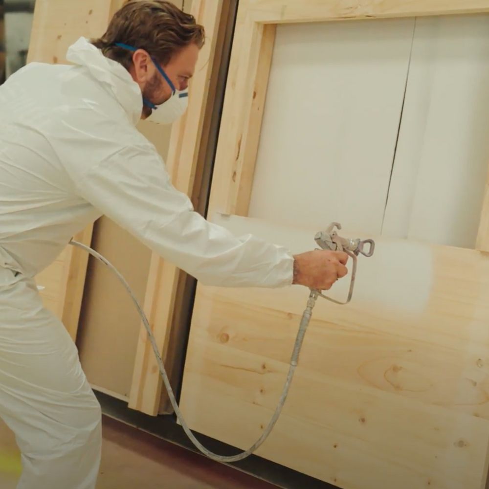A Castle & Cubby employee dresssed in a painters white uniform using a paint spray gun to paint a wooden cubby house wall in white.