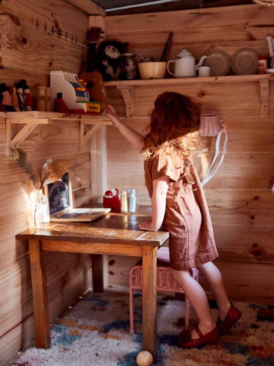 A red haired girl around 4 years of age inside her wooden cubby house, leaning over a table and reaching up to a shelf to her toys.