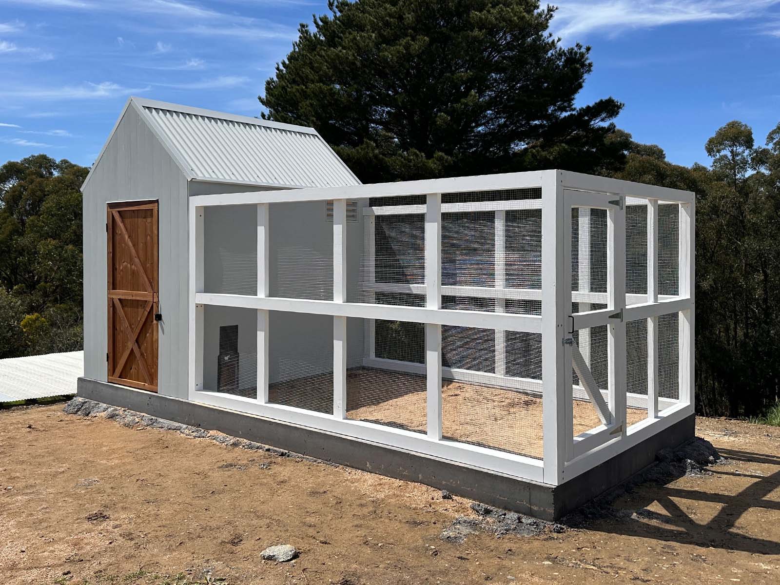 Large grey barn style chicken coop with white chicken run on a sunny day with trees in the background shot diagonally