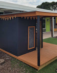 A commercial grade painted wooden cubby house with a wraparound verandah porch in a kindergarten playspace