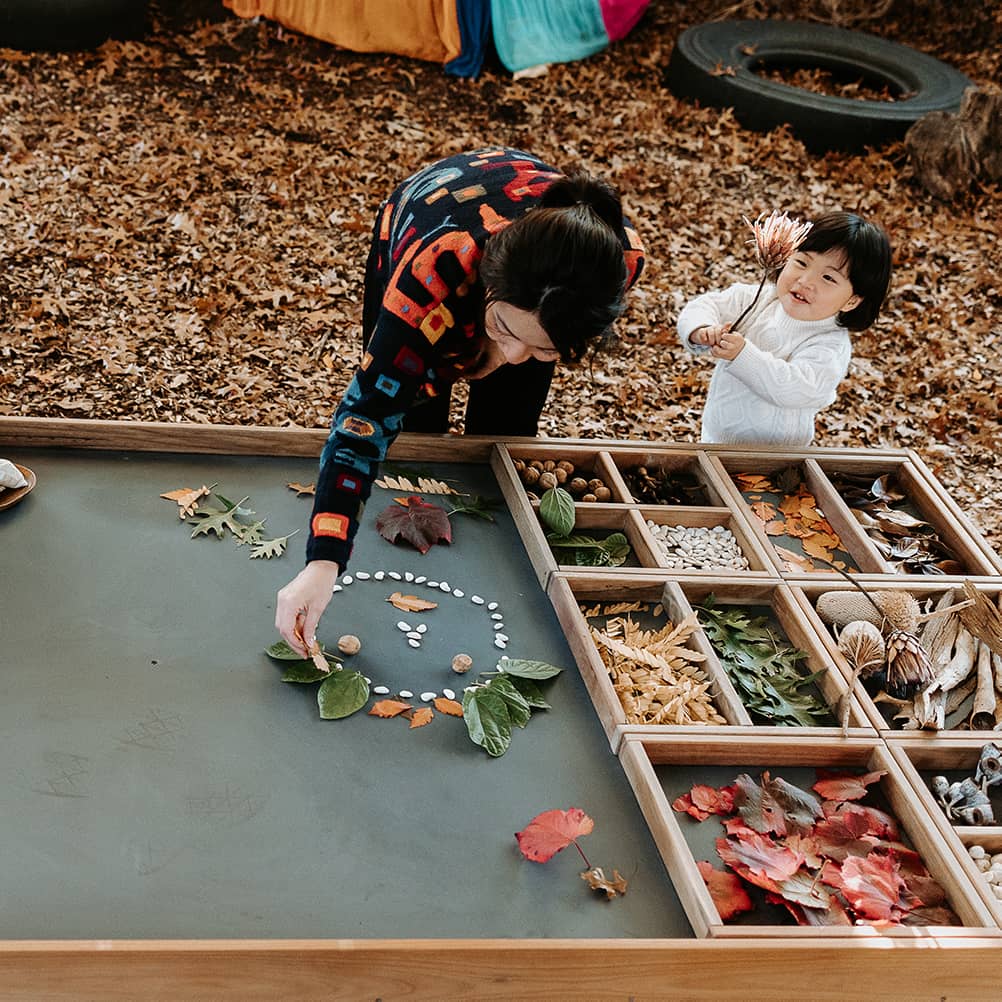 Educator and child creating a design on the chalkboard tabletop of an Exploration & Loose Parts Table in an outdoor ELC.
