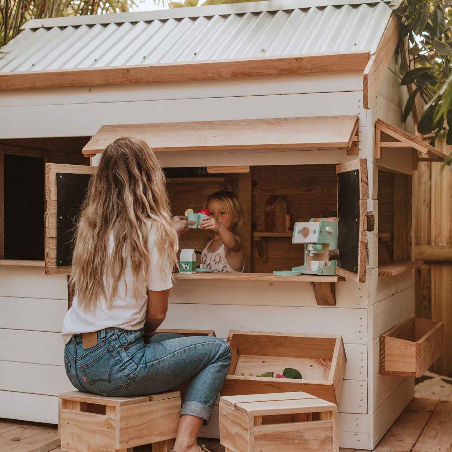 A mother & daughter interacting through a wooden cubby house window - they are clinking tea cups in celebration of their play time.