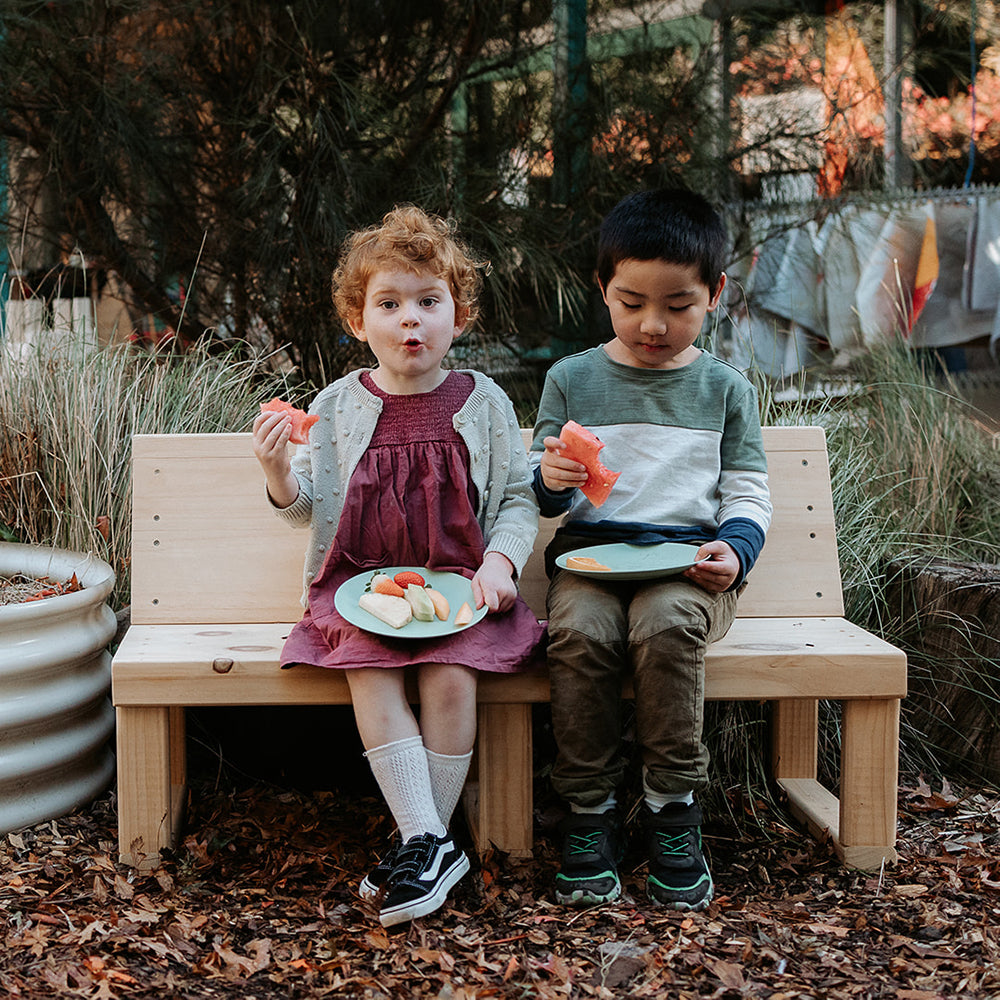 Two  kids sitting on a bench seat with back enjoying a fruit snack