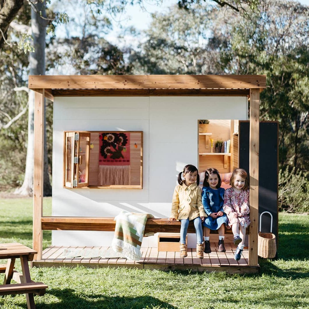 Three young girls sitting on the verandah of a modern wooden cubby house with flat roof