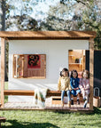 Three young girls sitting on the verandah of a modern wooden cubby house with flat roof