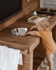 A baby plays at a beautifully crafted wooden outdoor mud kitchen bowel