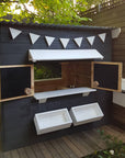 A close up view of the front of a painted timber cubby house with open windows and accessories