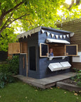 A painted wooden cubby house with a flat roof in a backyard under a big leafy tree