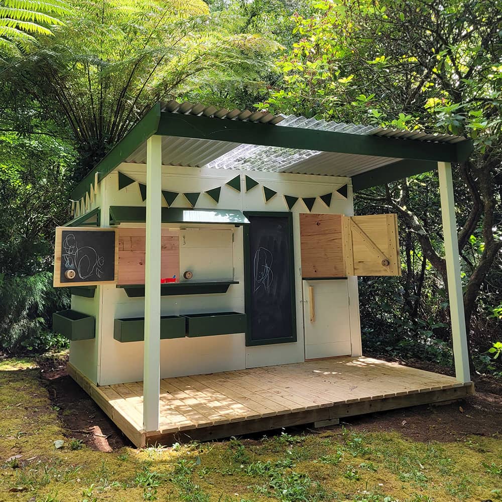 A wooden cubby house with a front porch verandah in dappled sunlight in a leafy garden