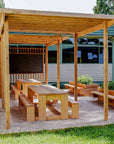Timber outdoor pergola with benches and tables under a wooden canopy in front of a building.