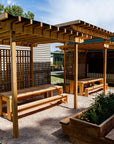 A timber shade structure with a mesh canopy and wooden benches underneath, situated in a school