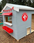 A grey painted wooden cubby house with red and white trims in a school play ground