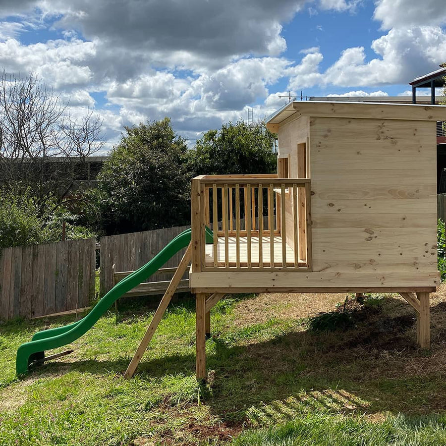 A side view of a large cubby house on a raised platform with a slide down the sloping grass