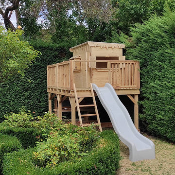 A large platform with handrail and cubby house and slide against a beautiful green hedge