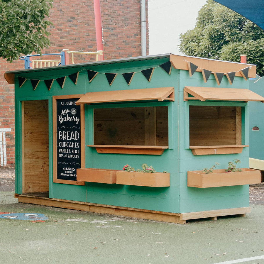 A green bakery cubby house in a school yard with chalkboard and timber accessories
