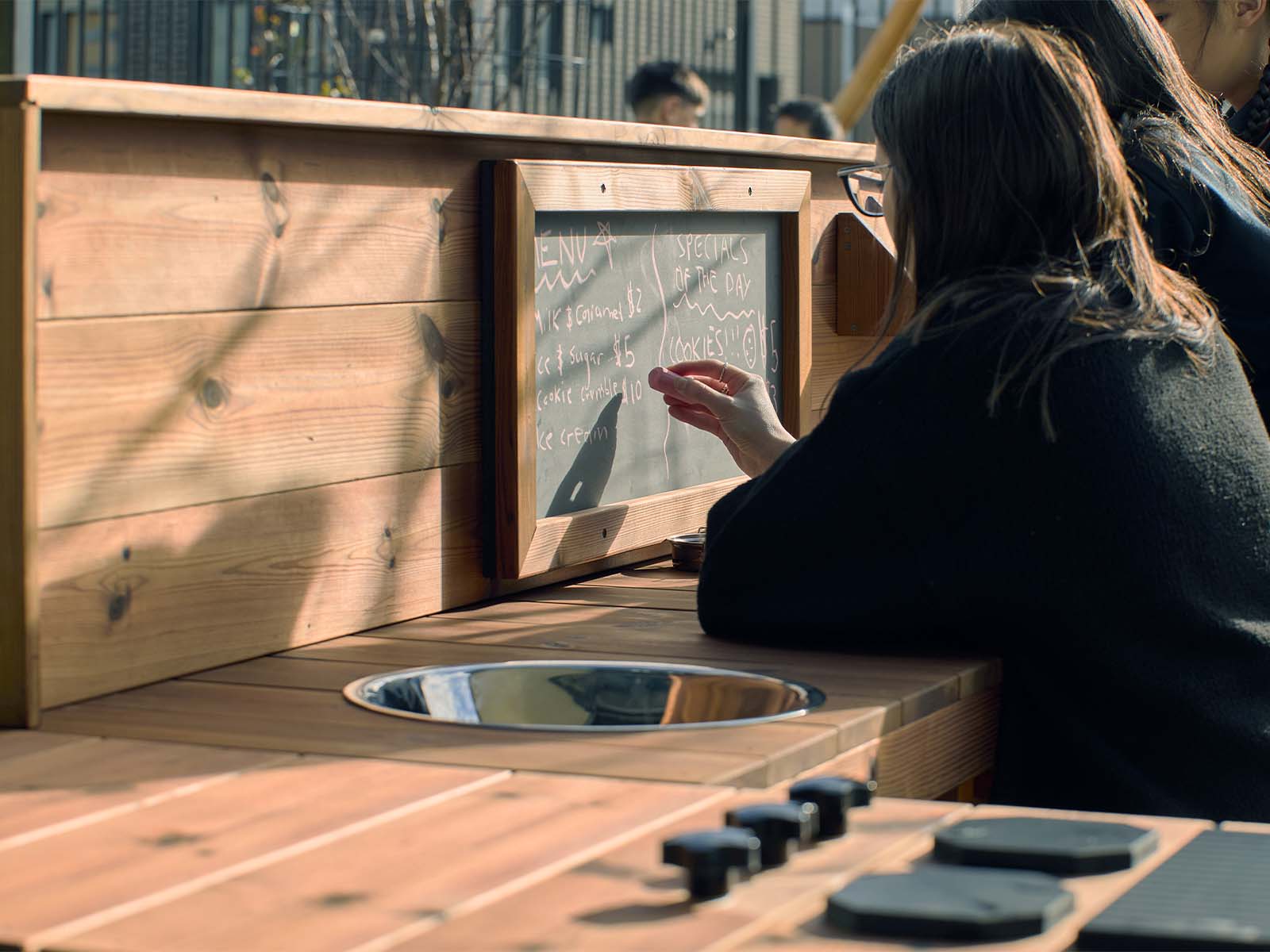 School teacher writing on wooden mud kitchen chalk board with sink, hotplates and knobs in foreground