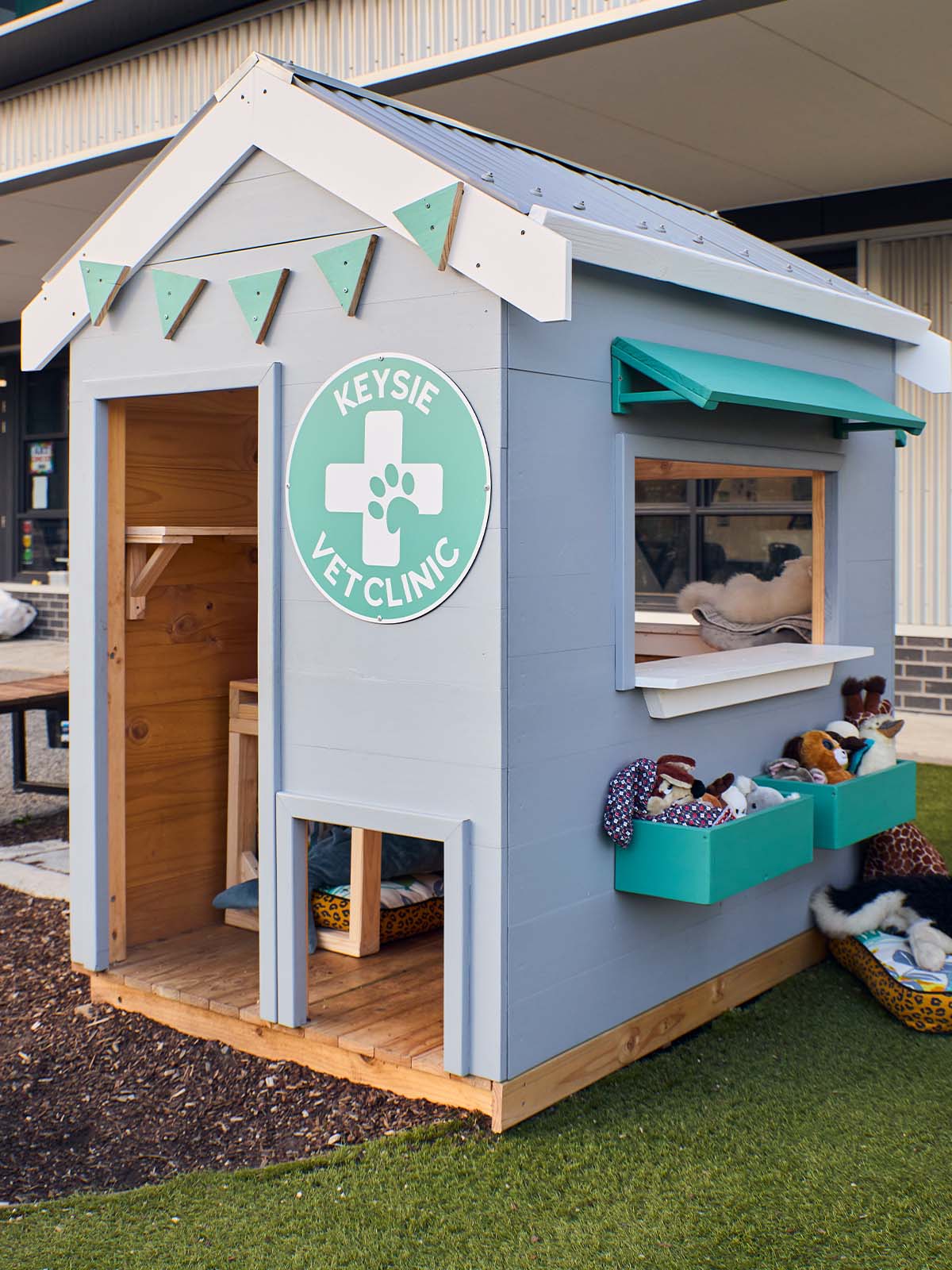 A vet themed grey and green cubby house in a school yard staged with soft toys 