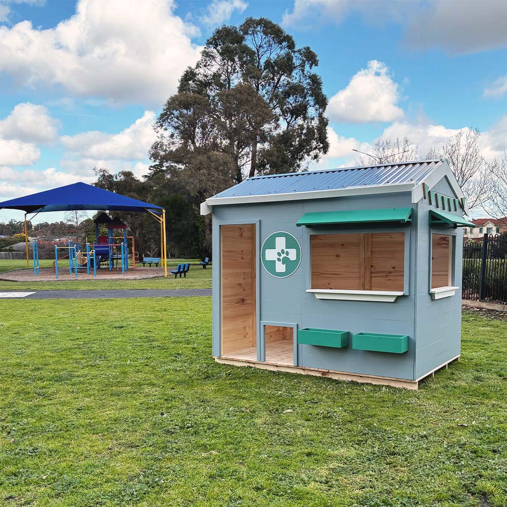 A vet themed grey and green cubby house on grass in a school yard with play equipment in the background