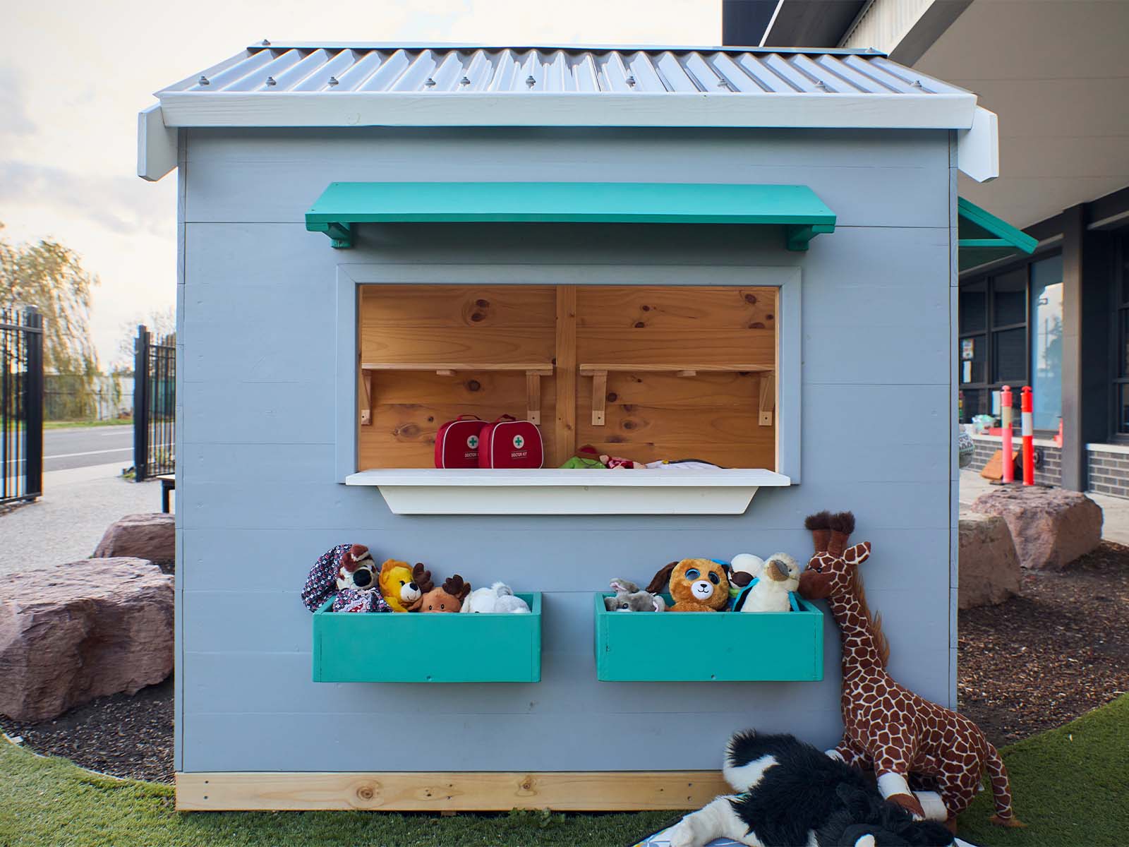 A vet themed grey and green cubby house shot from the side showing a large window in a school yard staged with soft toys 