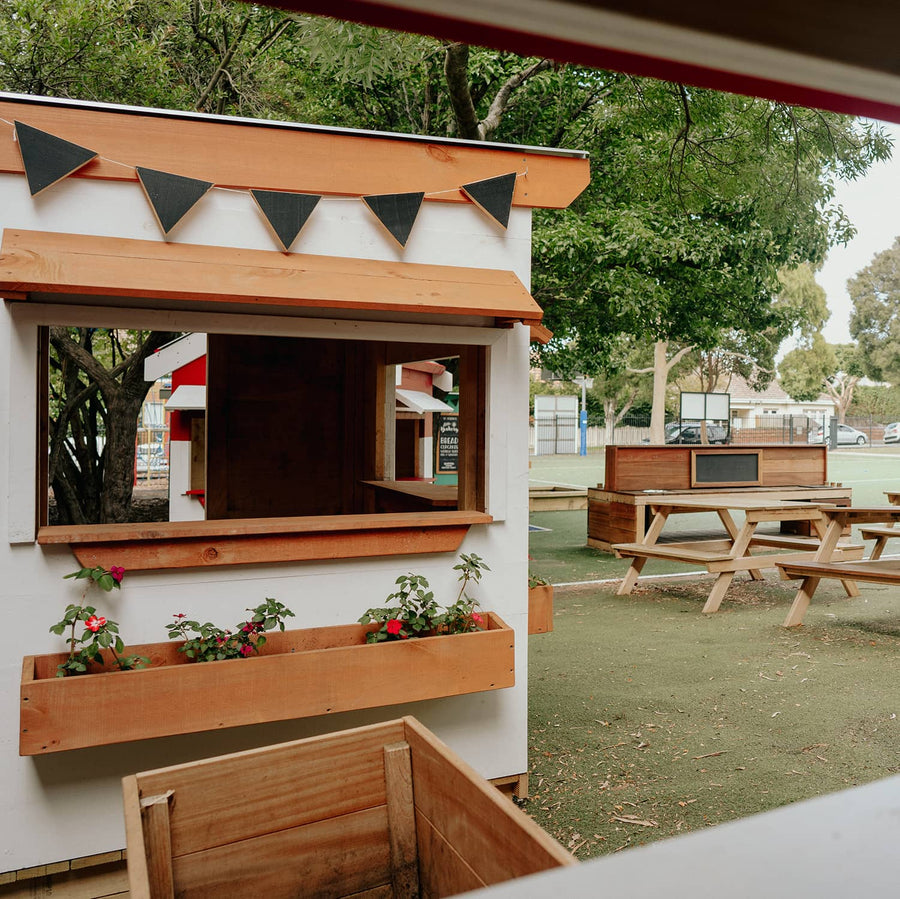 A white painted wooden cubby house on school grounds with a mud kitchen and picnic tables