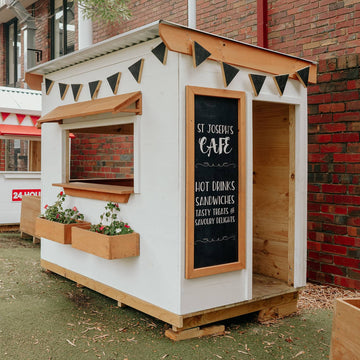 A bright white painted cafe cubby house with stained timber accents and chalkboard artwork in a school yard