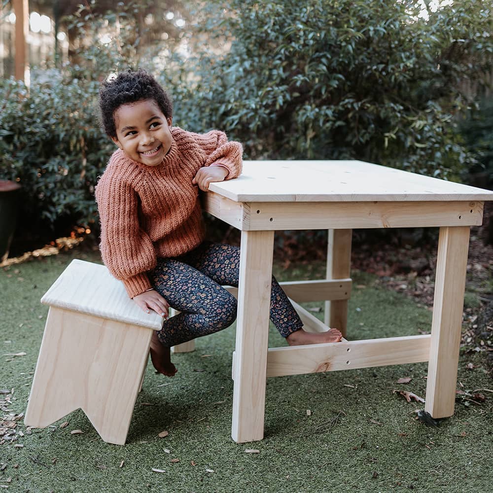 A girl sits at a table on a pine carpenter stool