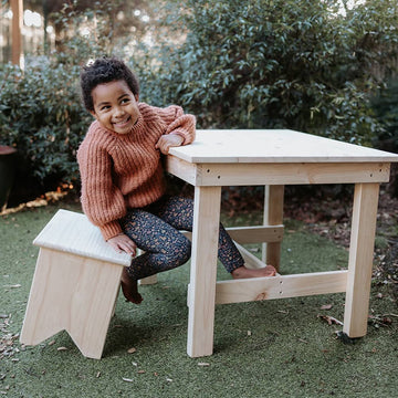 A girl sits at a table on a pine carpenter stool