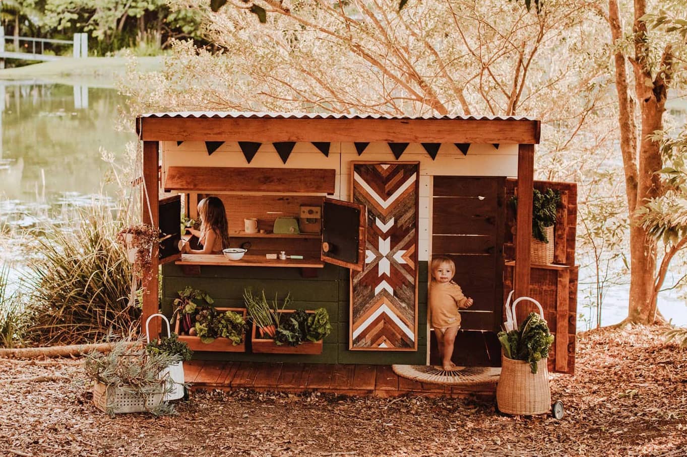 Castle and Cubby olive green and white cubby house with verandah. Positioned lakeside, a girl and small toddler play.