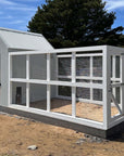 Large grey barn style chicken coop with white chicken run on a sunny day with trees in the background shot diagonally