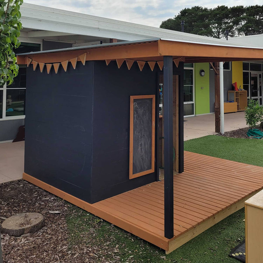 A commercial grade painted wooden cubby house with a wraparound verandah porch in a kindergarten playspace