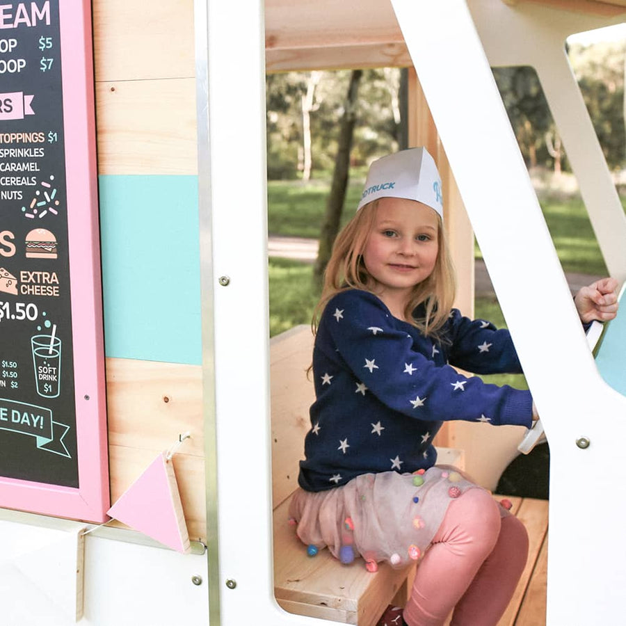 A girl sits in the drivers seat of a wooden foodtruck cubby house play space