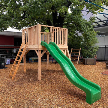 A sturdy wooden platform with handrail in a school yard with a bright green slide