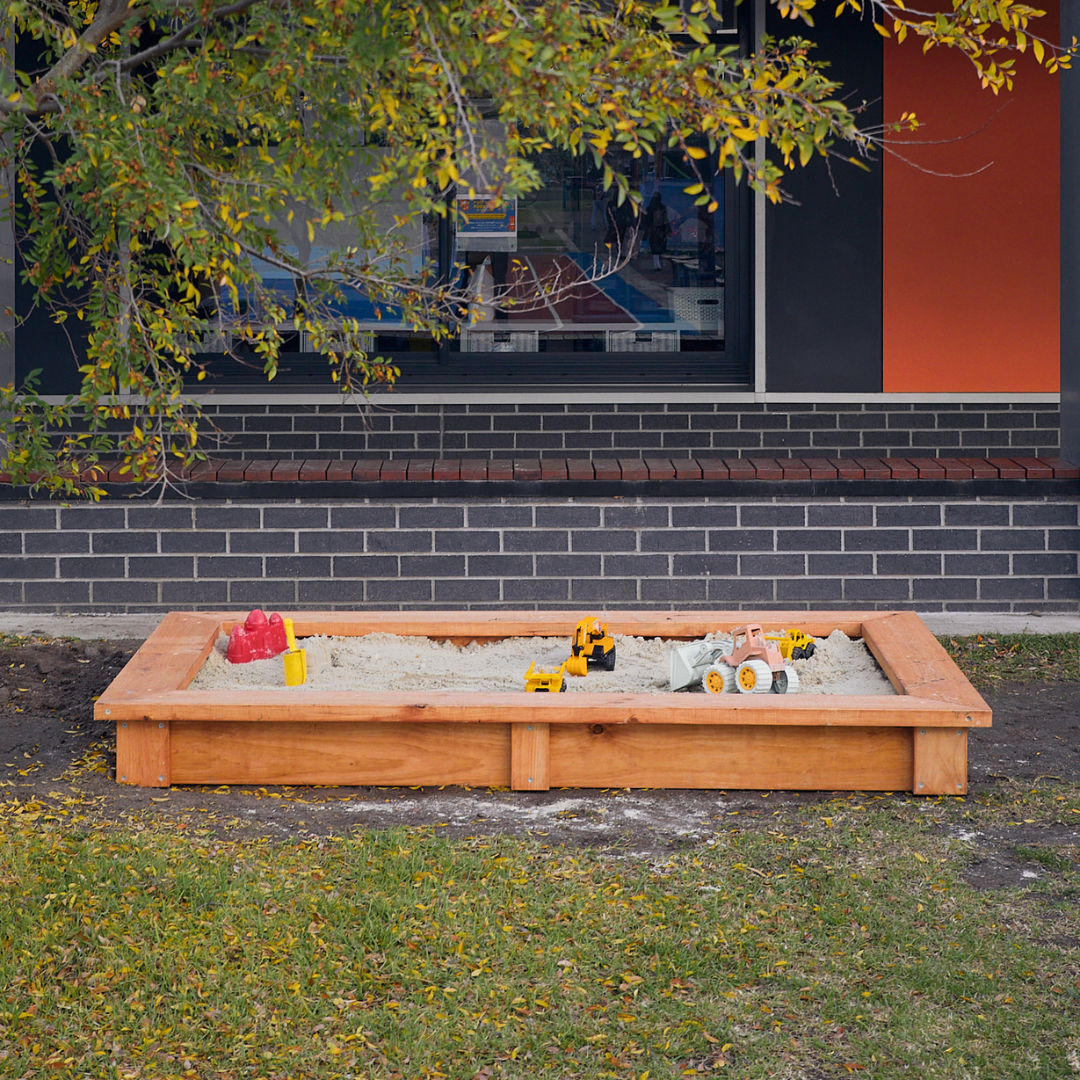 Raised timber sandpit in school grounds with toys and building in background
