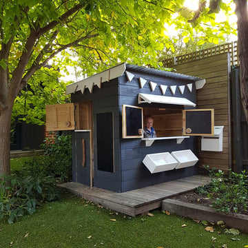 A painted wooden cubby house with a flat roof in a backyard under a big leafy tree