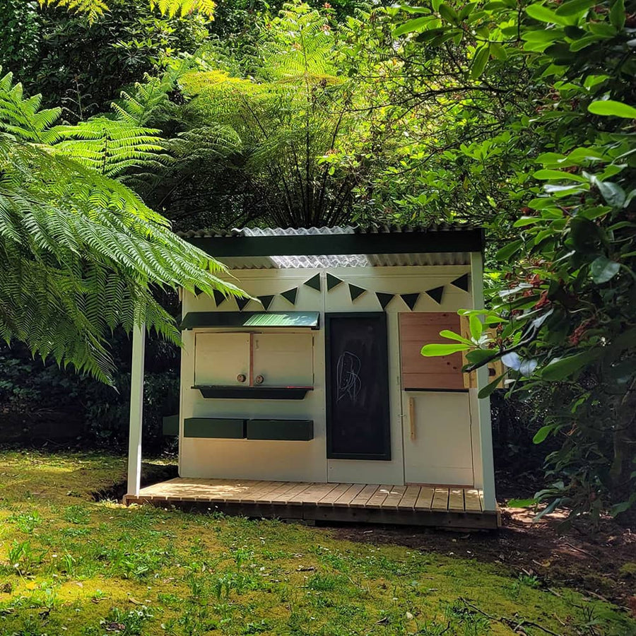A painted timber cubby house with a verandah in a backyard garden