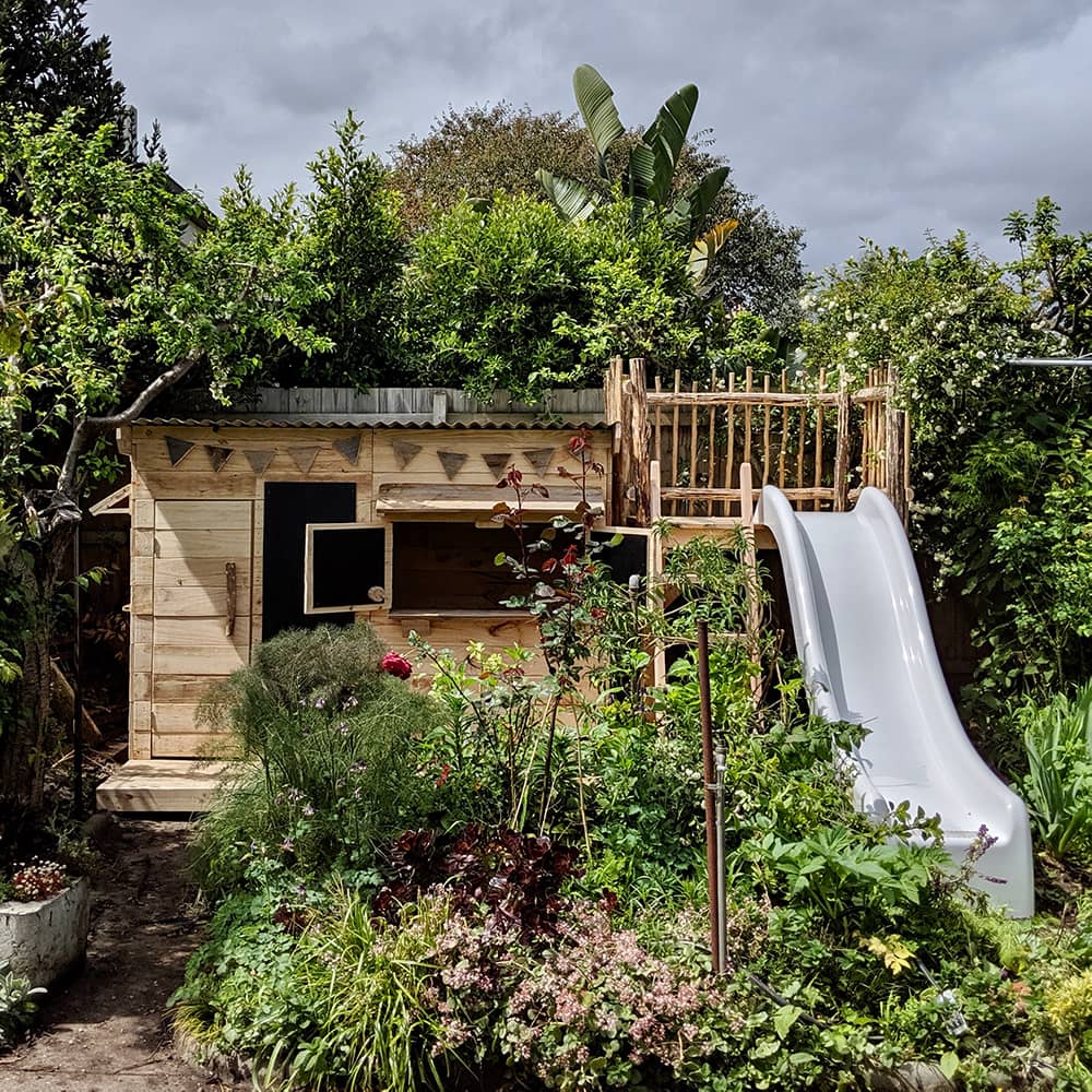 A cubby and wooden platform with slide are nestled into a beautiful overgrown garden