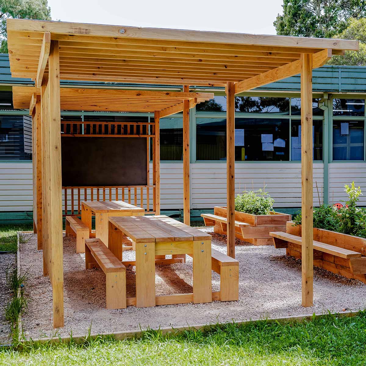 Timber outdoor pergola with benches and tables under a wooden canopy in front of a building.